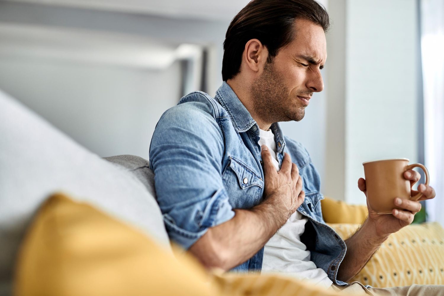 young-man-feeling-sick-holding-his-chest-pain-while-drinking-tea-living-room young-man-feeling-sick-holding-his-chest-pain-while-drinking-tea-living-room